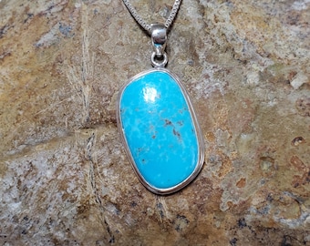 May include: A turquoise pendant necklace featuring a rectangular stone set in a silver-coloured metal frame. The bright blue stone has brown matrix patterns. The necklace chain is a delicate silver box chain. The pendant is displayed on a textured, brown stone surface.