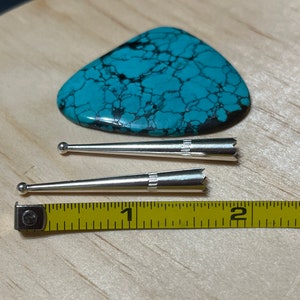 May include: Two silver, pointed, decorative pieces, each with a small ball at one end and a serrated edge at the other. The pieces are about 1 inch long and are lying on a wooden surface next to a yellow measuring tape.
