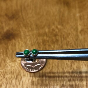 May include: A pair of green gemstone earrings held by tweezers over a US penny. The earrings are small and round, with a silver setting.