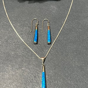 May include: A silver necklace and matching earrings with blue opal pendant and drop earrings. The pendant and earrings are long and narrow.