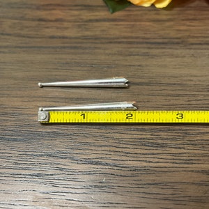 May include: Two silver metal knitting needles, one with a rounded tip and the other with a pointed tip, are placed on top of a yellow measuring tape. The tape measures in inches.