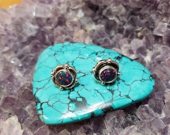 May include: A pair of silver stud earrings with round, iridescent opal gemstones set in a silver bezel. The earrings are displayed on a turquoise and purple rock.