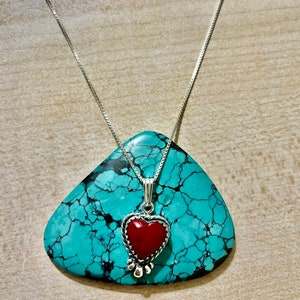 May include: A silver chain necklace with a red heart pendant. The pendant is set in a silver frame with a detailed design. The necklace is displayed on a turquoise stone.