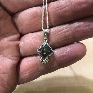 May include: A silver chain necklace with a square-shaped pendant featuring a black opal gemstone. The pendant has a silver border and three silver balls hanging from the bottom.