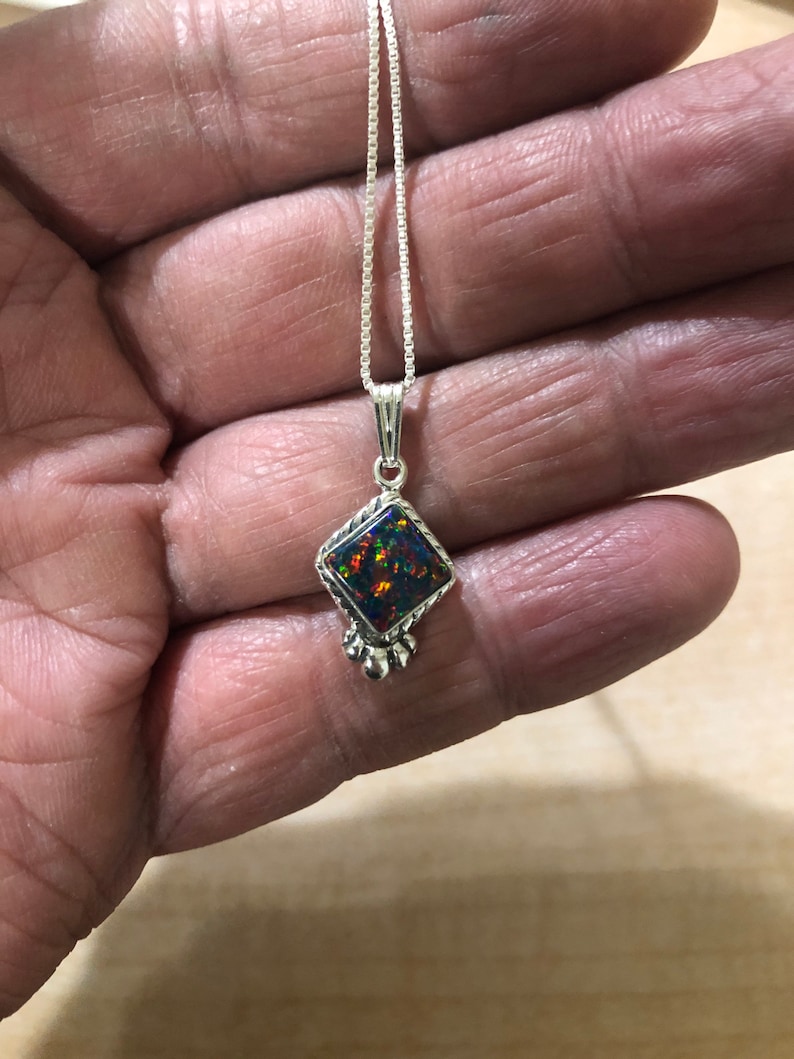 May include: A silver chain necklace with a square-shaped pendant featuring a black opal gemstone. The pendant has a silver border and three silver balls hanging from the bottom.