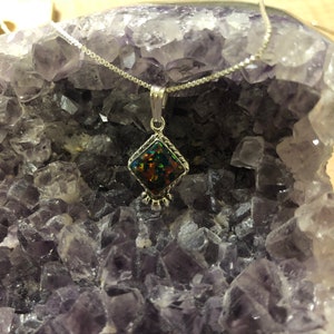 May include: A silver chain necklace with a pendant featuring a dark iridescent gemstone set in a silver diamond-shaped frame. The necklace is displayed on a bed of purple amethyst crystals.