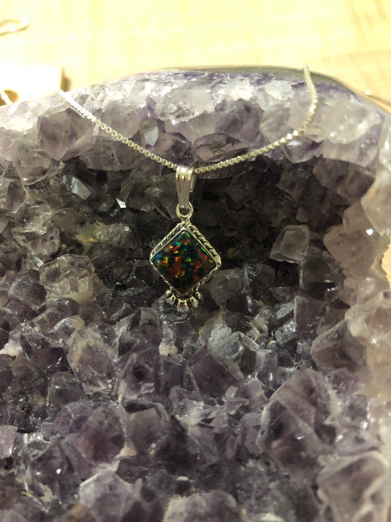 May include: A silver chain necklace with a pendant featuring a dark iridescent gemstone set in a silver diamond-shaped frame. The necklace is displayed on a bed of purple amethyst crystals.