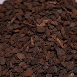 May include: Close-up of a pile of dark brown, irregularly shaped pieces. The pieces appear to be small, flat, and textured, with varying shades of brown and hints of lighter tones. The overall texture is rough and organic.