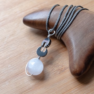 Può includere: Una collana d'argento con un ciondolo in pietra di luna bianca e un ciondolo in argento con un design geometrico. La collana è su un cordoncino grigio.