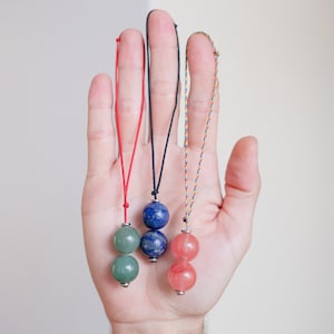 May include: Three necklaces with different coloured cords and gemstone beads. The first necklace has a red cord and two green round beads. The second necklace has a black cord and two blue round beads. The third necklace has a multi-coloured cord and two pink round beads.
