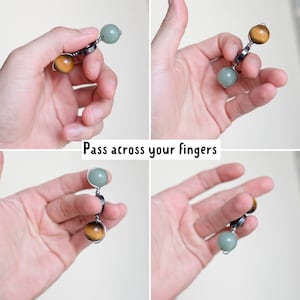 May include: A hand holding a beaded fidget toy with green and brown beads. The beads are connected by silver wire and a silver ring. The text "Pass across your fingers" is visible below the hand.