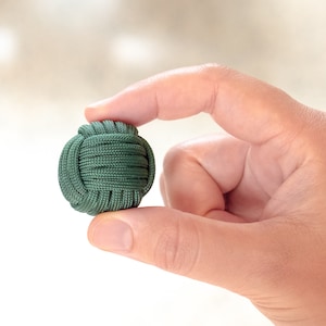Puede incluir: Una bola de paracord verde, también conocida como puño de mono, se sostiene en la mano de una persona. La bola está hecha de paracord verde tejido apretado.