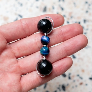 Op de afbeelding: Een handgemaakte hanger met een verticale opstelling van gepolijste kralen. Het ontwerp omvat twee grote, glanzende zwarte kralen aan de boven- en onderkant, met twee kleinere, levendige blauwe en turquoise kralen in het midden, allemaal verbonden door zilverdraad.