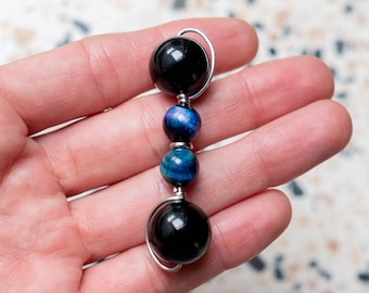 May include: A handcrafted pendant featuring a vertical arrangement of polished beads. The design includes two large, glossy black beads at the top and bottom, with two smaller, vibrant blue and teal beads in the centre, all connected by silver wire.