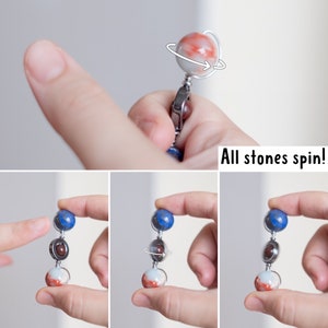 May include: A spinning, multi-colored, gemstone necklace with a silver chain. The necklace features three round stones: a blue lapis lazuli, a red jasper, and a white agate. The stones are connected by silver rings and spin freely. "All stones spin!" is written below the necklace.