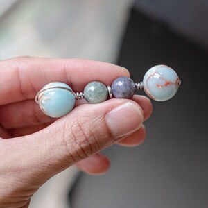 May include: A close-up of a hand holding a wire-wrapped beaded bracelet. The bracelet features four beads: a light blue bead, a green bead, a purple bead, and a light blue bead with brown streaks.