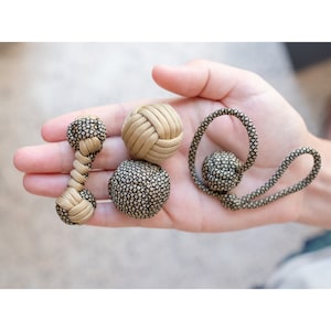 Pode incluir: Uma coleção de nós decorativos em várias formas. Os nós são feitos de cordão bege e preto, com uma variedade de padrões. Um tem a forma de um haltere, outro é esférico e outro é um laço, todos segurados em uma mão.