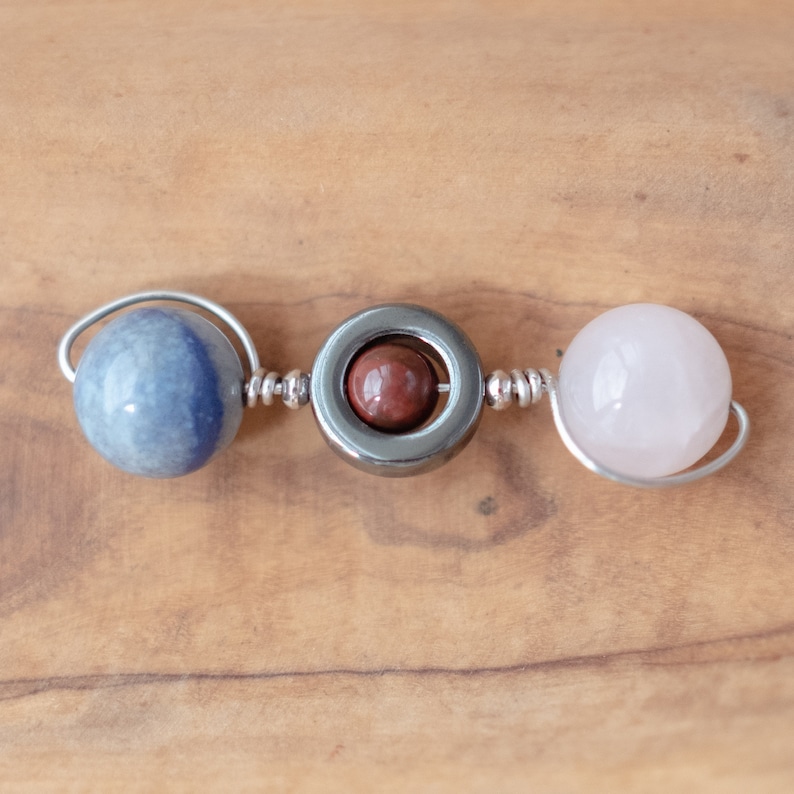 May include: A silver wire wrapped bracelet with three beads: a blue agate, a hematite bead with a red jasper stone inside, and a pink rose quartz bead.