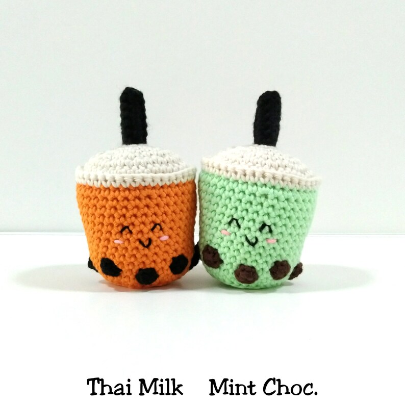 May include: Two crocheted boba tea plushies, one orange with black boba pearls and the other green with brown boba pearls. Both have white tops and black straws. The orange one says "Thai Milk" and the green one says "Mint Choc."