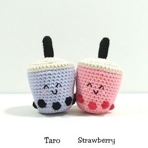 May include: Two crocheted boba tea cups, one purple with black boba pearls and one pink with red boba pearls. The cups have smiling faces and are labeled "Taro" and "Strawberry".