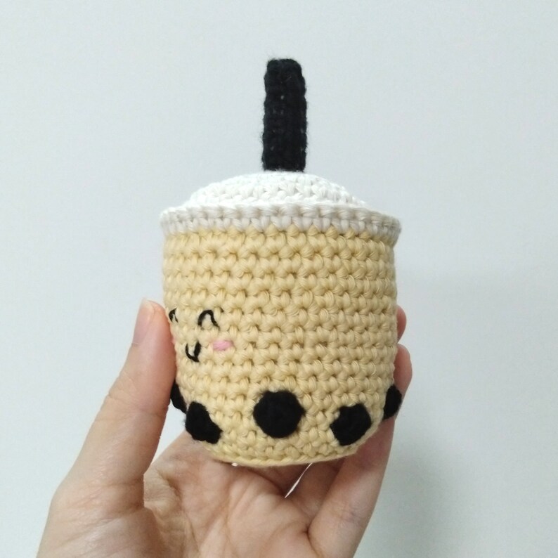 May include: A crocheted boba tea cup with a black straw and a white lid. The cup is beige with black boba pearls and a cute pink and black face.