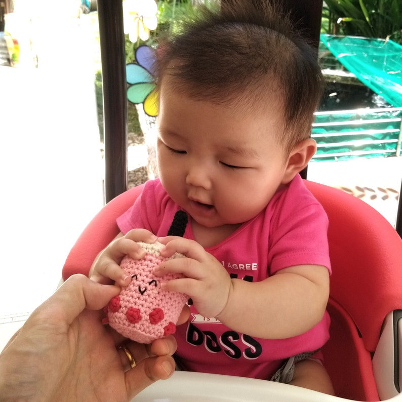 May include: A young child in a pink shirt with the word "Boss" on it is holding a pink and white crocheted toy. The toy has a smiling face and pink polka dots.