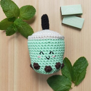 May include: A crocheted green and white boba tea cup with a brown straw and brown boba pearls. The cup has a smiling face with pink cheeks. The cup is surrounded by green mint leaves.