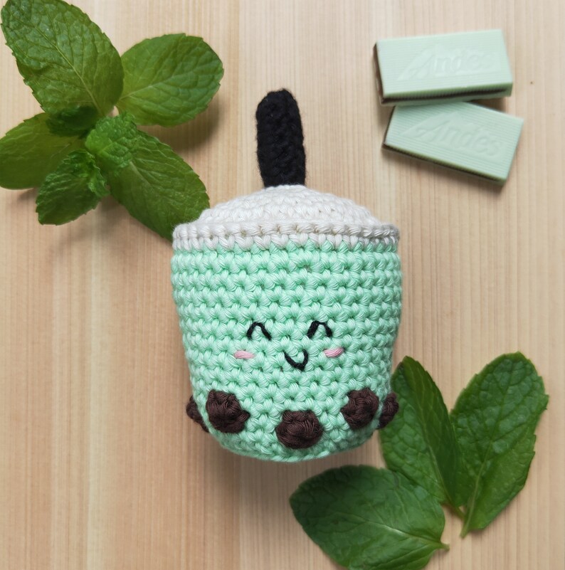 May include: A crocheted green and white boba tea cup with a brown straw and brown boba pearls. The cup has a smiling face with pink cheeks. The cup is surrounded by green mint leaves.