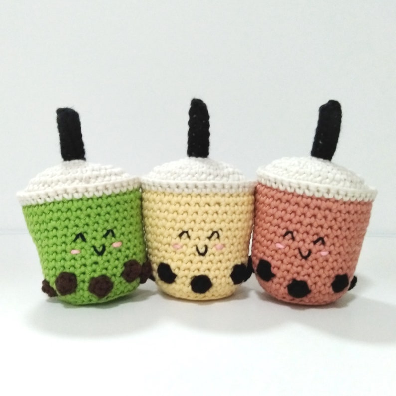 May include: Three crocheted boba tea cups with smiling faces. The cups are green, yellow, and pink. Each cup has black boba pearls at the bottom.