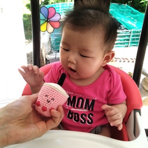 May include: A pink crocheted boba tea cup with a smiling face, held by a person. A young child wearing a pink shirt with the text "Mom is the Boss" is looking at the cup.