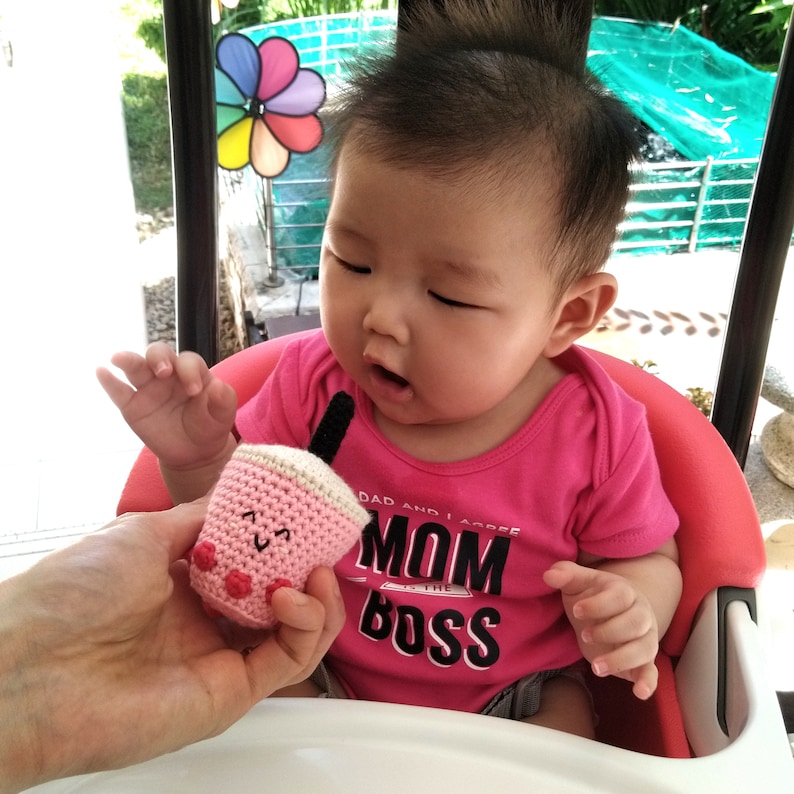May include: A pink crocheted boba tea cup with a smiling face, held by a person. A young child wearing a pink shirt with the text "Mom is the Boss" is looking at the cup.