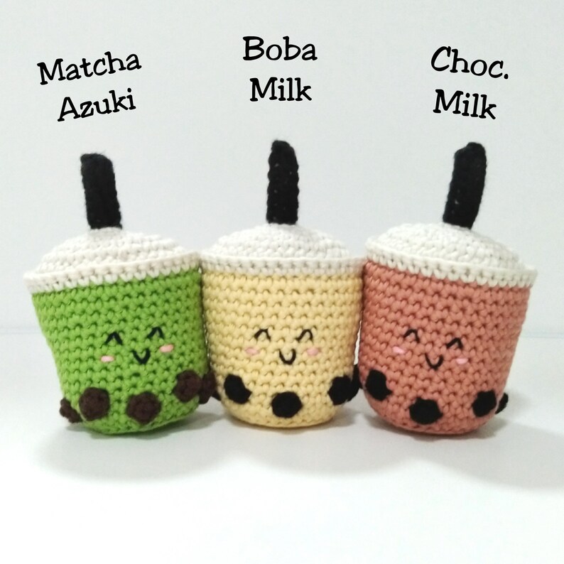 May include: Three crocheted boba tea cups with smiling faces. The cups are green, yellow, and pink, and each has black boba pearls at the bottom. The text above the cups reads "Matcha Azuki", "Boba Milk", and "Choc. Milk".
