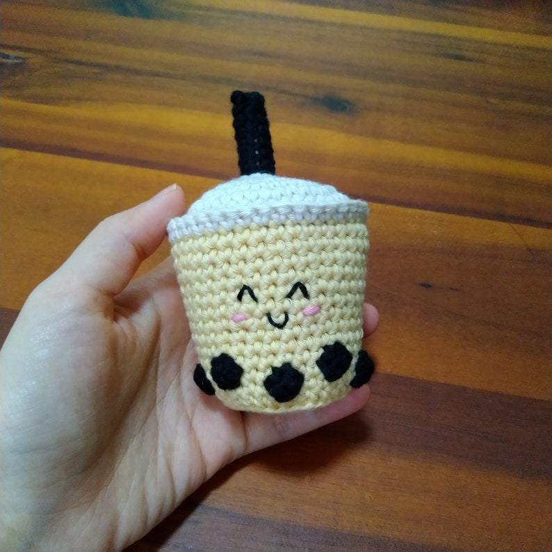 May include: A crocheted boba tea cup with a smiling face and black boba pearls. The cup is yellow with a white lid and a black straw.