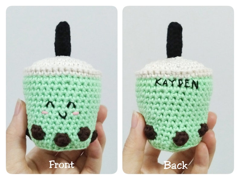 May include: A green crocheted boba tea cup with a black straw and brown boba pearls. The cup has a white crocheted lid and a smiling face. The back of the cup has the name "KAYDEN" embroidered on it.