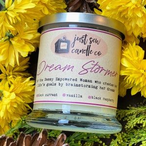 May include: A glass candle jar with a brown label that reads "just sew candle co" and "Dream Stormer" in pink lettering. The label also includes the text "for the Sassy Empowered Woman who creates her life's goals by brainstorming her dreams" and lists the scents "black currant", "vanilla", and "black raspberry".