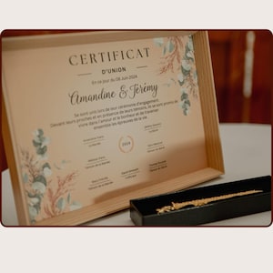 Può includere: Un certificato incorniciato con il testo "CERTIFICAT D'UNION" e i nomi "Amandine & Jérémy". Il certificato è in una cornice di legno chiaro, con un oggetto color oro in una scatola nera in primo piano. Il certificato è datato 8 giugno 2024.