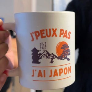 May include: White ceramic mug with the text "J'PEUX PAS J'AI JAPON" in orange. The mug features a graphic of a pagoda, mountains, a sun, and a bonsai tree. The mug has a curved handle.