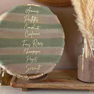 Peut inclure: Un élément décoratif rond avec des rayures alternées de tissu vert sauge et beige. Des lettres dorées en script épellent des mots comme "Amour" et "Confiance". La pièce repose sur un support doré et un plateau en bois, avec un vase en verre de fleurs séchées à proximité.