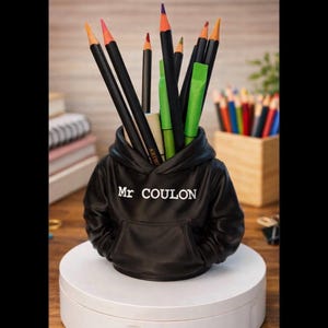 Peut inclure: Un porte-crayons noir en forme de mini sweat à capuche, avec le texte "Mr COULON" en blanc. Il contient des crayons de couleur et des stylos verts. Le support est sur une plateforme circulaire blanche.