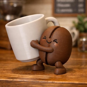 May include: A whimsical figurine of a smiling coffee bean character hugging a white coffee mug. The coffee bean is brown with a happy face and small legs. The mug is white and has a handle. The figurine is on a wooden surface.