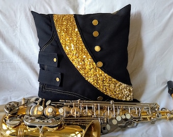 Puede incluir: Un cojín decorativo negro con detalles de lentejuelas doradas y botones dorados. El cojín tiene un diseño de estilo militar y está colocado frente a un saxofón dorado.