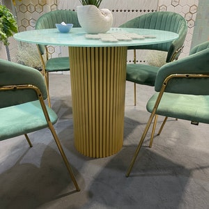 May include: A round glass table with a gold metal base and four teal velvet chairs. The table has a light blue glass top and a gold metal base with a fluted design. The chairs have a teal velvet upholstery and gold metal legs.