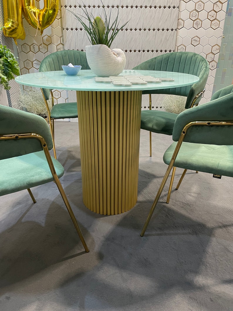 May include: A round glass table with a gold metal base and four teal velvet chairs. The table has a light blue glass top and a gold metal base with a fluted design. The chairs have a teal velvet upholstery and gold metal legs.