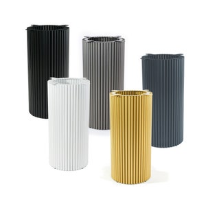 Op de afbeelding: Vijf cilindrische metalen plantenbakken in verschillende kleuren: zwart, grijs, wit, goud en donkergrijs. De plantenbakken hebben een geribbeld ontwerp en zijn in een rij gerangschikt.