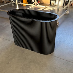 Peut inclure: Un bac à plantes rectangulaire noir avec des rainures verticales. Le bac a un bord supérieur arrondi et est fabriqué dans un matériau sombre. Le design est moderne et minimaliste, adapté à l'intérieur ou à l'extérieur. Hauteur d'environ 100 cm.