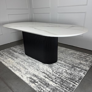 Könnte beinhalten: Runder weißer Esstisch mit einem schwarzen, geriffelten Untergestell. Der Tisch steht auf einem grauen und weißen Teppich mit Muster.