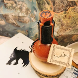 Może przedstawiać: Pomarańczowa butelka z miksturą z czarną szyjką i emblematem wilka na korku. Butelka jest oznaczona napisem "Swallow" i stoi na drewnianym plastrze. Mapa i książka z grafiką wilka znajdują się w tle.