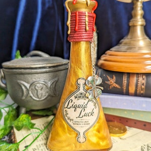 Può includere: Una bottiglia di pozione dorata con l'etichetta "Liquid Luck" e un ciondolo a forma di quadrifoglio. La bottiglia ha un sigillo di cera rosso e oro ed è riempita con un liquido scintillante. Un calderone e monete d'oro sullo sfondo.