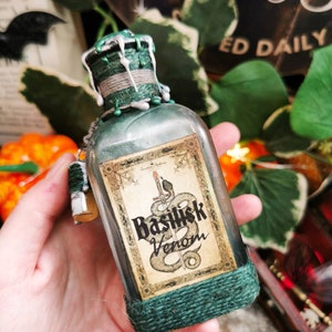 Puede incluir: Una botella de vidrio de estilo vintage con una etiqueta que dice "Basilisk Venom". La botella está envuelta en hilo verde y tiene una tapa decorativa con cera verde y detalles plateados. Un pequeño dije cuelga de la botella.