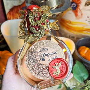Könnte beinhalten: Eine runde Glasflasche mit der Aufschrift "Phoenix Tears" mit einer Phönix-Illustration und einem roten Wachssiegel. Die Flasche ist mit goldfarbenen Blättern und einem roten Stoffaufsatz verziert. Der Inhalt der Flasche ist eine hell bernsteinfarbene Flüssigkeit.